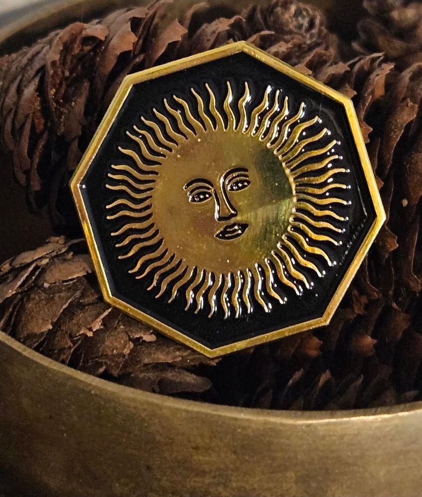 A photo of an enamel pin in a golden bowl of brown pine-cones. The pin has an octagonal shape and a golden frame with a golden sun, with a face, in the center. The gold is against a black base.