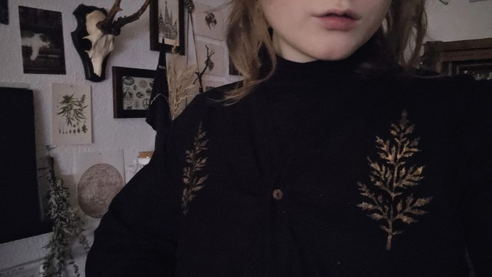 Pictute of my chest, wearing a black dress with golden Mugwort blockprint in the shoulder area on both sides, and a black crew neck. Behind me is a wall filled with art and postcards depicting nature motifs, as well as a dried mugwort and an old antlered deer skull mount.