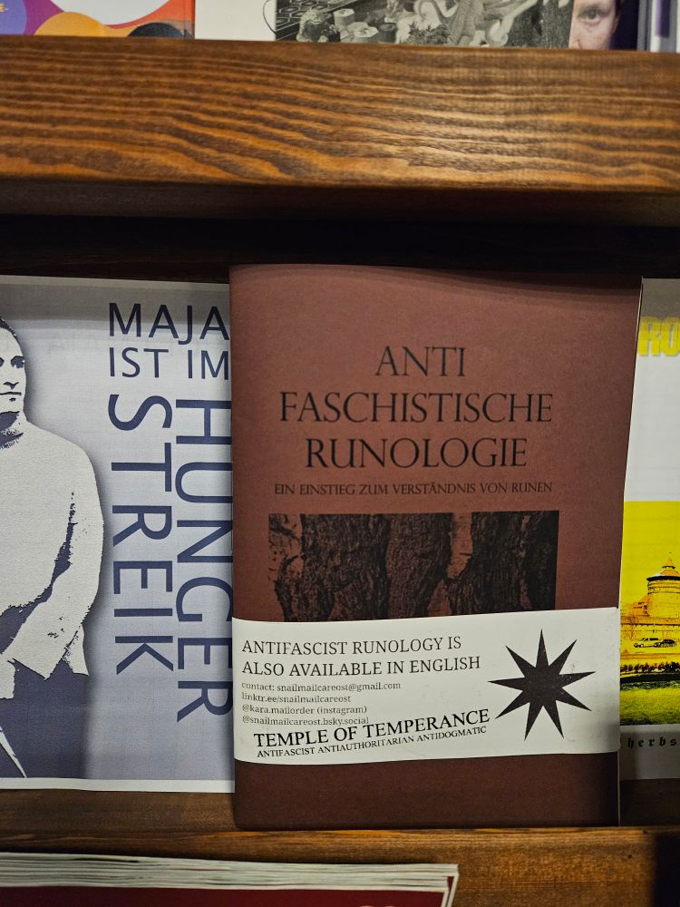 Mein Zine "Antifaschistische Runologie, Ein Einstieg Zum Verständnis Von Runen" in brauner Farbe mit einer weißen Banderole auf der steht "Antifascist Runology is also available in english" darunter meine Kontaktdaten. Das Zine liegt zwischen Flyern.