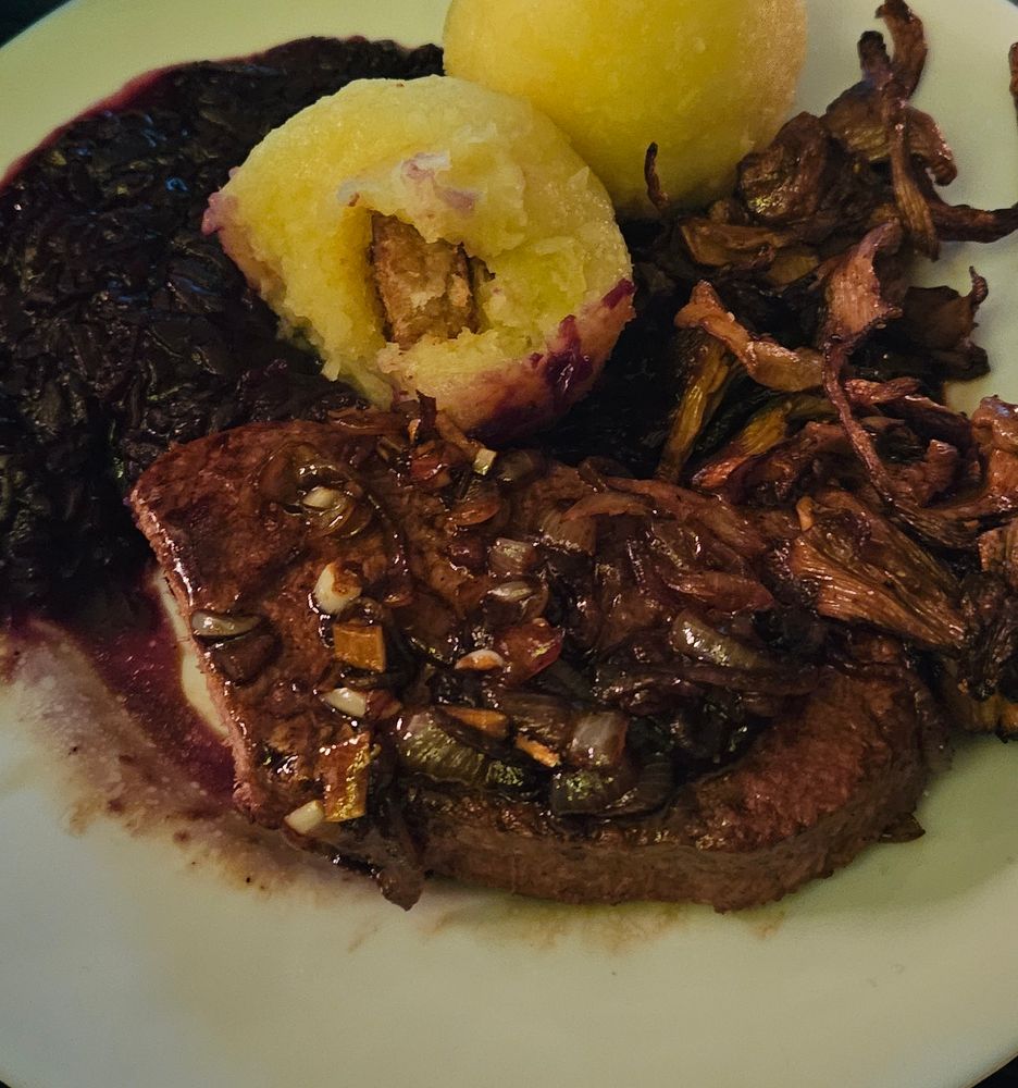 Foto eines Tellers mit Klößen, kross gebratenen Pfifferlingen, dunklem Rotkraut, und einem dunklen Veganen Steak getoppt mit ein paar dunklen Zwiebeln und Knoblauch. Es sieht alles sehr saftig und gesättigt aus und die Klöße stoßen golden hervor. Das Steak und der Kloß sind angeschnitten. Im inneren des Kloßes kommt ein Crouton hervor.