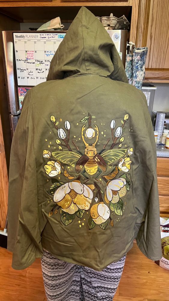 Green (military green ish) cloak with embroidered white and gold flowers with thick petals and a large golden beetle with two long horns (I don’t know the species, sorry!!). The cloak is about hip length and viewed from the back. 