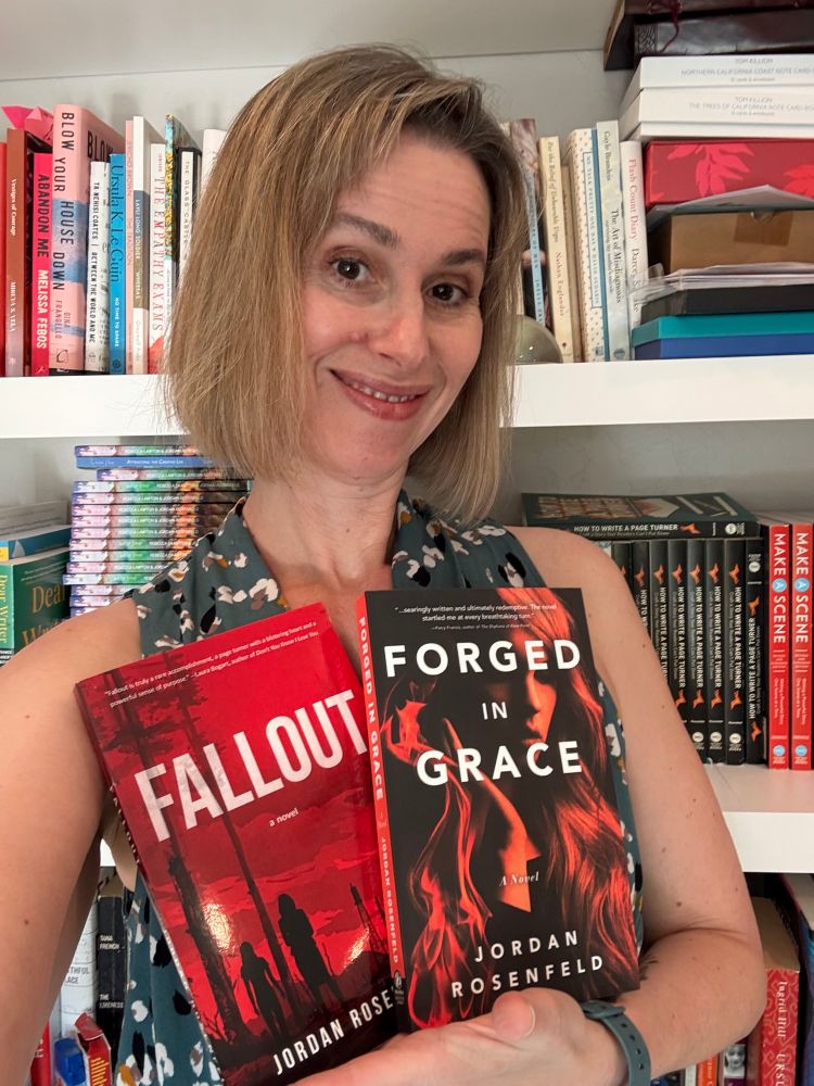 A photo of Jordan, a white woman with a dark blonde bob, holding her two novels Fallout (a red cover with the silhouette of two women in a forest) and Forged in Grace (a woman’s face veiled in flames) against the backdrop of Jordan’s bookshelf. 