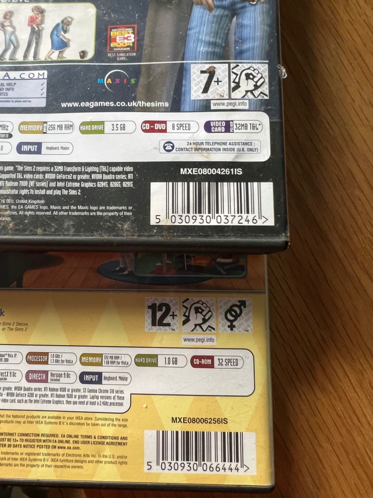 A photo showing the back of two video game boxes, but only the bottom corner of each box, showing the age rating, age rating descriptors, some of the recommended specs and the barcode. 

The first box is rated PEGI 7 and belongs to The Sims 2. It has the symbol for “violence” next to the age rating. 

The second box is rated PEGI 12 and belongs to The Sims 2 IKEA Home Stuff. It has the symbol for “violence” and “sex references” next to the age rating