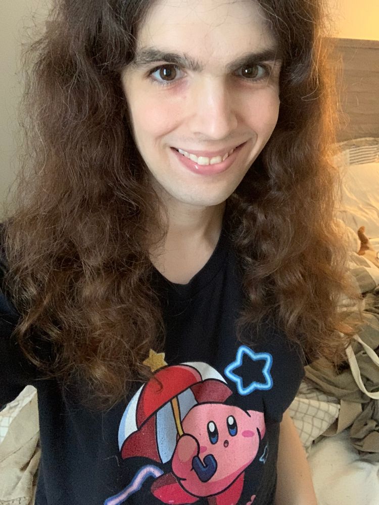 Mari in 2025, with natural curly hair, wearing t-shirt of Kirby holding a parasol.