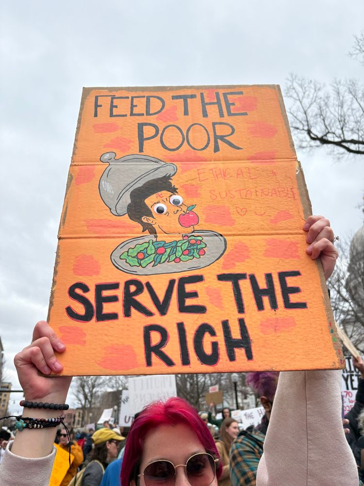 photo of someone with red/pink hair holding up a sign that says “feed the poor, serve the rich” with a cartoon drawing of elon musk’s head (with googly eyes) on a platter with an apple in his mouth
