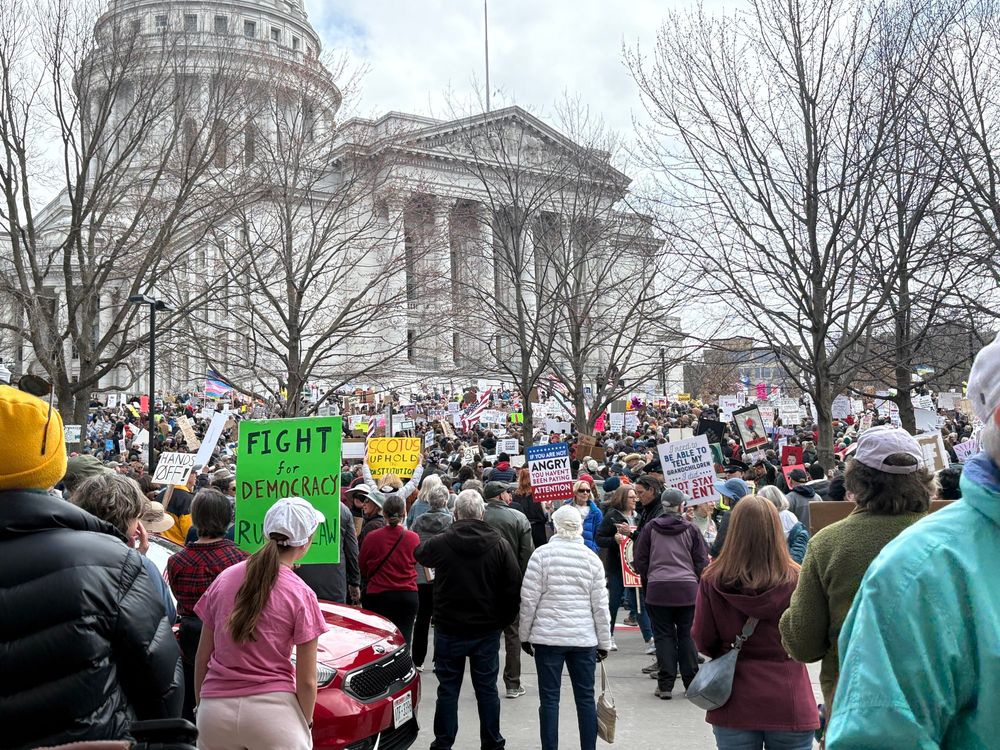 photo of a large crowd of people, many carrying signs, standing in front of the Madison WI capitol building 