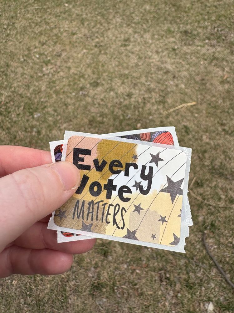 photo of me holding two stickers in my hands above a grass background. The sticker on top says “every vote matters” with some stars on it and colors brown, yellow, and pinkish 