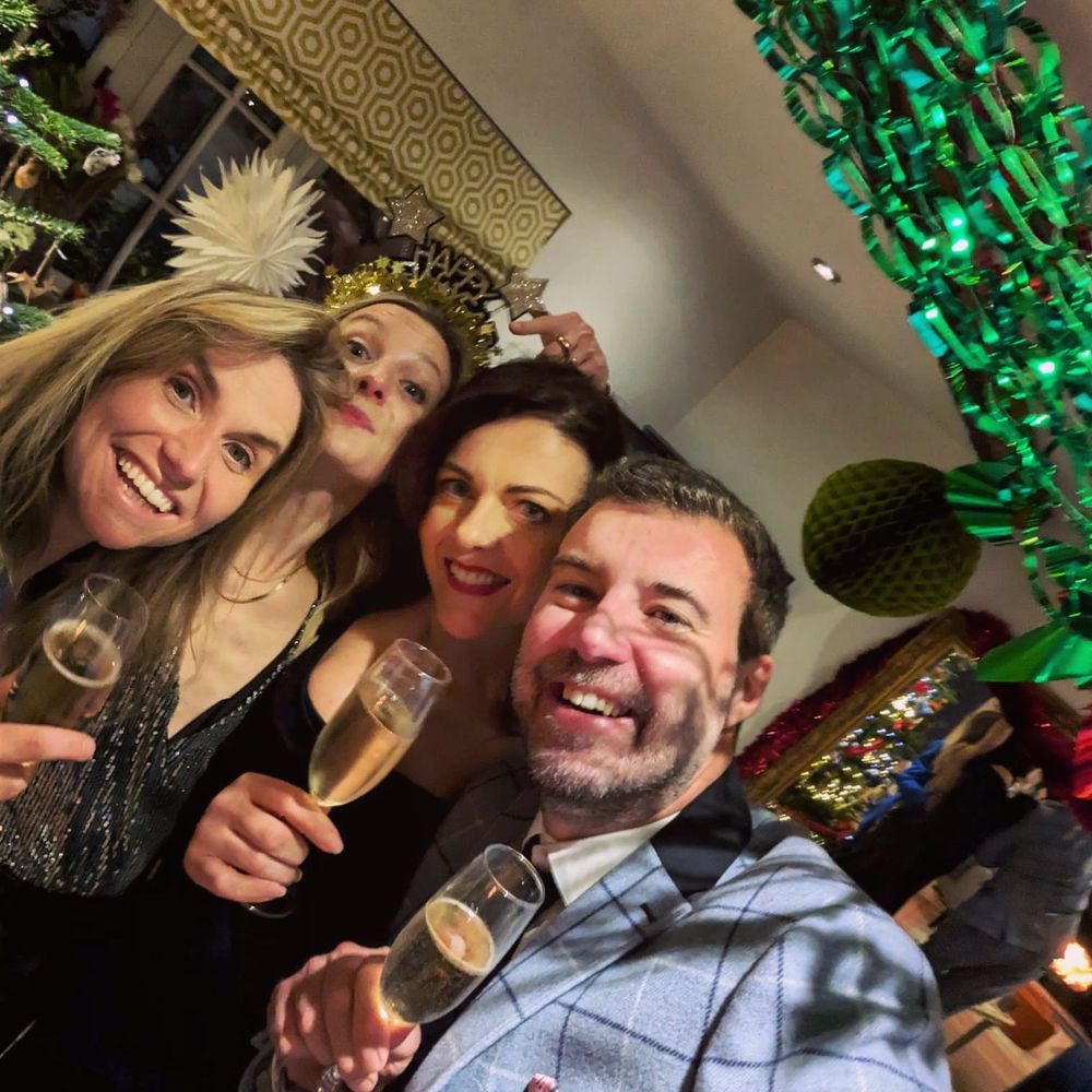 Four people smile to camera, three are holding champagne flutes, it is NYE.