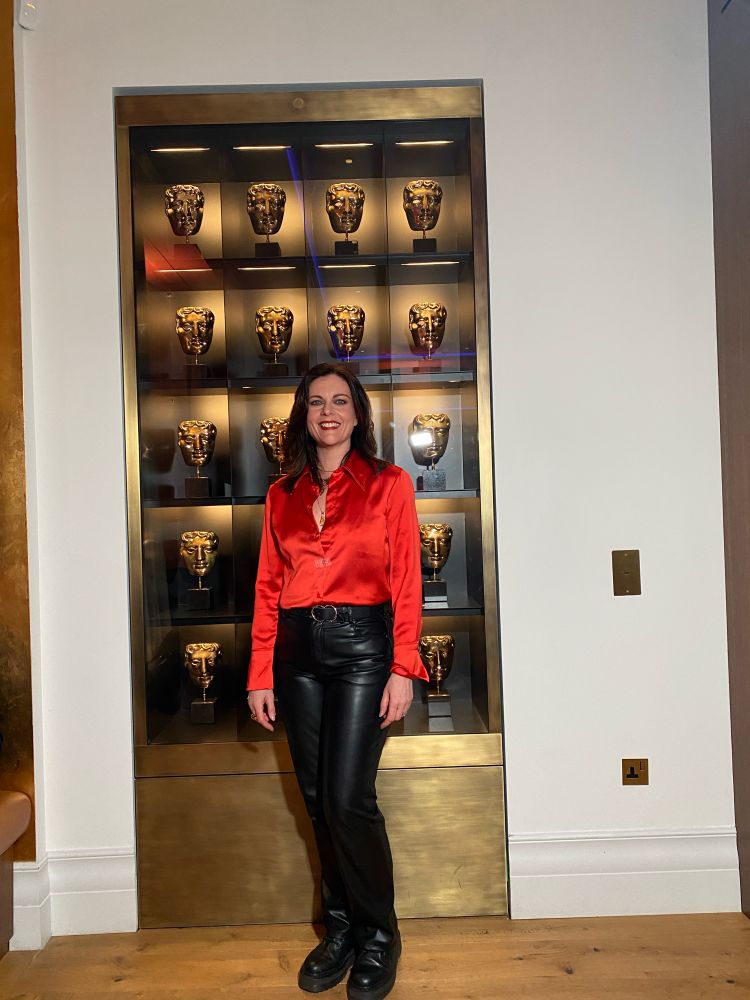 A woman in a deep orange satin shirt and leather trousers stands infront of a cabinet full of BAFTAS