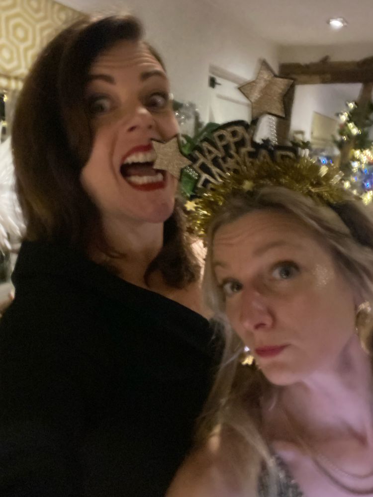 Two ladies face the camera. The shorter blond lady on the right is wearing a ‘Happy New Year’ headress. The taller dark haired lady is pretending to bite one of the headdresses stars.