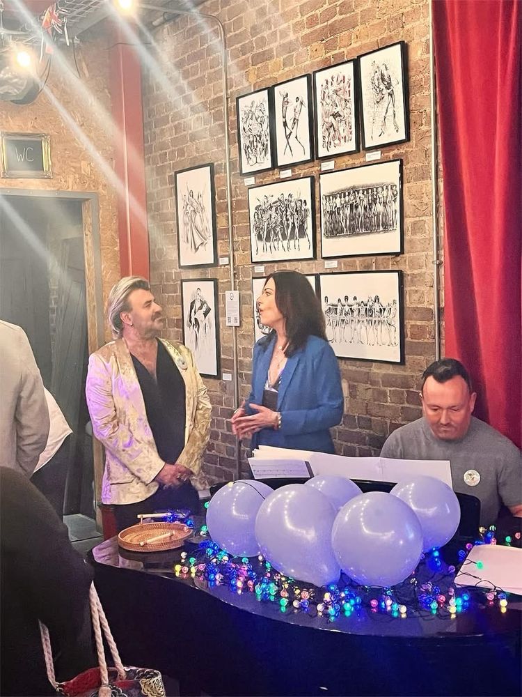 A man in a gold jacket and a woman in a blue suit stands behind a piano, the woman is singing at the man. The pianist is staring intently at the music as he plays. There are blue balloons and multicoloured string lights on the piano