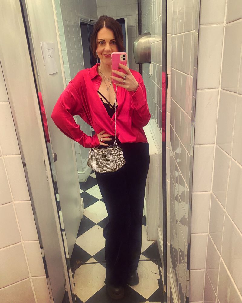 A lady in a pink shirt, black trousers and a silver sparkly crossbody bag stands with one hand on her hip whilst taking a full length selfie in a mirror. She is in the loo of a pub the walks are white tiles, the floor is black and white diamond checkerboard 