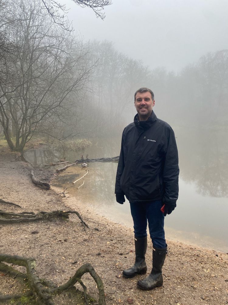 A smiling man in wellies, jeans, blue coat and black gloves stands in front of a pond in a forest. It is very misty.
