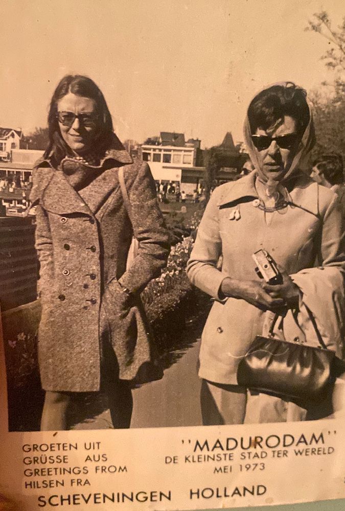A mother and daughter are strolling along a street in Holland. The daughter, in the left, has shoulder length brown hair and has her hands in her coat pockets a bag is slung over her left shoulder. The mum has dark hair, is wearing a head scarf and holding a camera and a handbag- a coat is slung over her left arm. Both are wearing sunglasses 