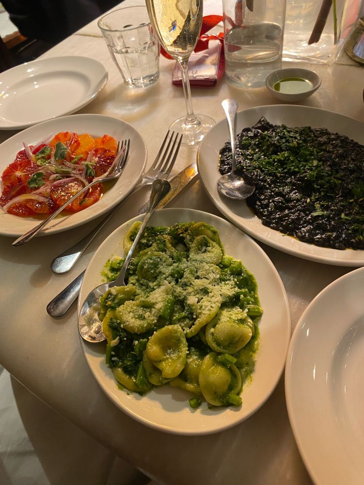 Three plates of delicious Italian food. Top left blood orange salad, top right black squid ink and cuttlefish risotto and bottom middle a green plate of paccheri with pummarola 