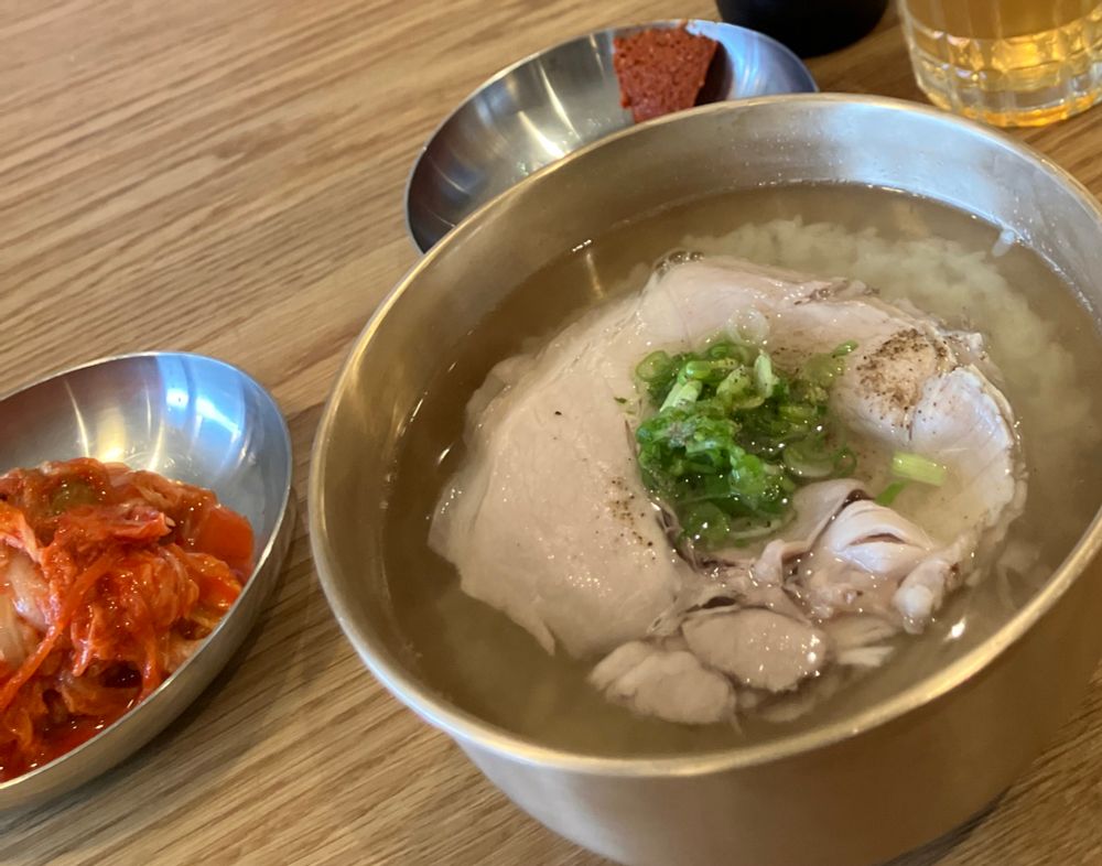 A bowl of pork broth with slices of pork, rice, and thin diced scallions.  Also featured, a small bowl of kimchi and a small bowl of fermented red paste. 