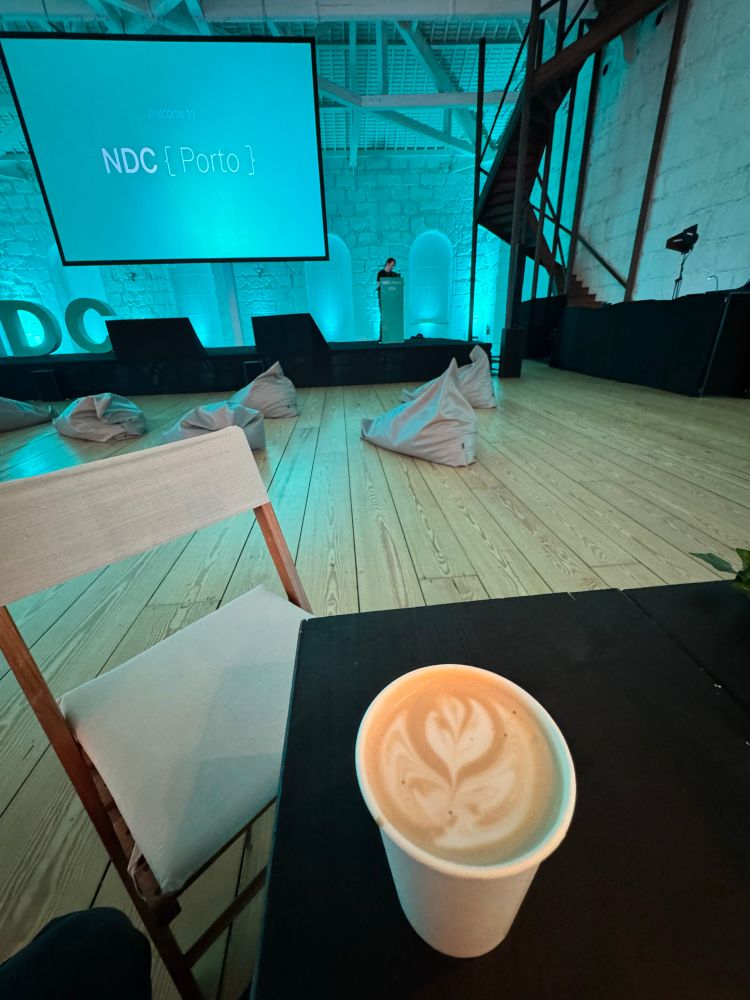Cappuccino with flower art in foreground 
Dylan Beattie on stage preparing 
Screen behind reads welcome to NDC Porto
