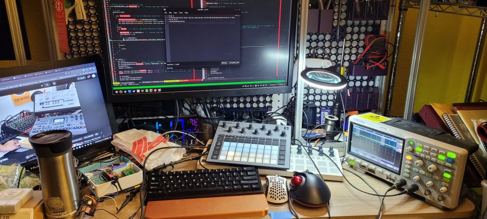 Desk covered in 2 synthesizers, a diy guitar pedal with circuitry exposed, an oscilloscope showing the midi signal, and a computer screen with C++ code and a serial terminal printing garbage.