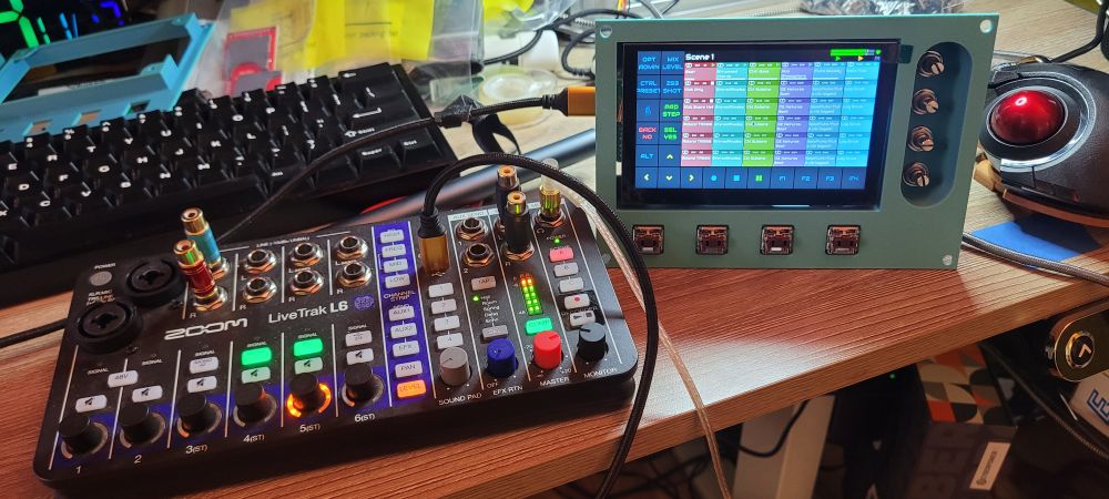Photo of a portable 6 inputnmixing board and a teal plastic 3D printed control panel with a 5 inch touch screen taking up the majority of nit, 4 mechanical keyboard buttons along the bottom, and 4 recessed rotary encoders, missing knobs, down the right side of the panel 