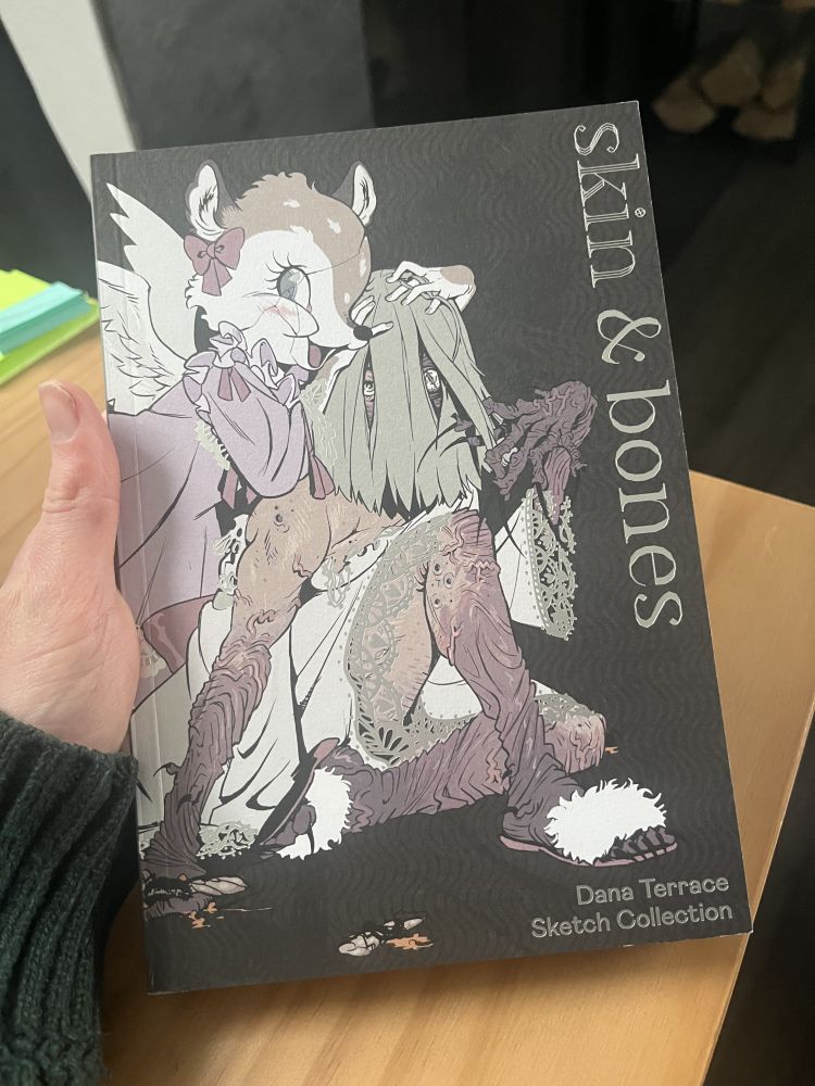 A copy of the book "skin & bones", a sketch collection from the artist Dana Terrace. 

The book cover features a macabre illustration of porcelain doll with angel wings, a faded lavender dress, and a deer's head with a crack running down its face grinning, one eye looking towards the viewer.

Within the illustration, said creature is holding the head of a human figure with long faded green hair strands obscuring its face, revealing only its eyes, one of that of a human and another of that of a feline with a gash in its iris, looking out towards the viewer.

The human figure is wearing open-toed thin sandals with a white fuzzy fur atop the sandal's strap and the body is adorned in a white dress with lace hems, printed in a silvery reflective paper, revealing the figures arms and legs

Those arms and legs become darker, more wrinkled, and veiny as they extend away from the torso and to the hands or feet. The hand of the human figure evokes an ancient decrepit corpse with long, curved, and pointed black fingernails.

The whole illustration is set on a greyish-black background with a subtle pattern of vertical waves comprising its texture. To the right of the illustration, in silvery reflective paper, reads the text "skin & bones" shown in a serif typeface, aligned sideways with the tops of each letter being close to the edge of the cover. 

In the bottom right corner, also printed in that reflective paper, the text "Dana Terrace" is displayed with the words "Sketch Collection" on bottom in a normal text alignment. 