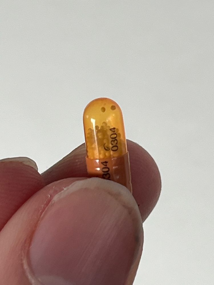 One orange 10mg capsule-pill of Dexedrine being held upright by a finger and a thumb. 

The capsule’s bottom half is opaque and orange-colored while the top half is a translucent yellowish-orange color which reveals the inside of the capsule, displaying the medication. 

This medication in the form of tiny white balls that can move freely within the capsule and are stacked close to the top but not all the way.

There are two tiny white balls sticking near to the top of the capsule’s interior and appear like two cute little eyes, therefore making the Dexedrine appear like a little guy.
