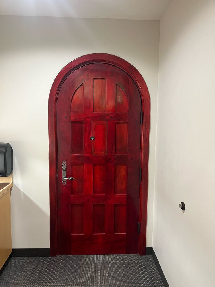 A red-stained wooden door with its top in the shape a rounded half-circle. The door is embossed to create a gridded pattern evocative of medieval gates. The door handle on the left is a grey polished metal with a handle that swirls.

In the top center of the door, is a smaller peakhole door in the same shape as the main door with a sliding lock that can open.

The entire door is oddly placed too, being tucked away in a nook in a normal, mundane conference room apart of a library.

Photo taken at Beacon Hill Library in Seattle, Washington, United States of America, in November 2024.
