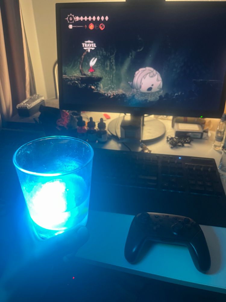 Photo of Bee holding a glowing glass of cyan liquid while playing hollow knight: silksong in the background