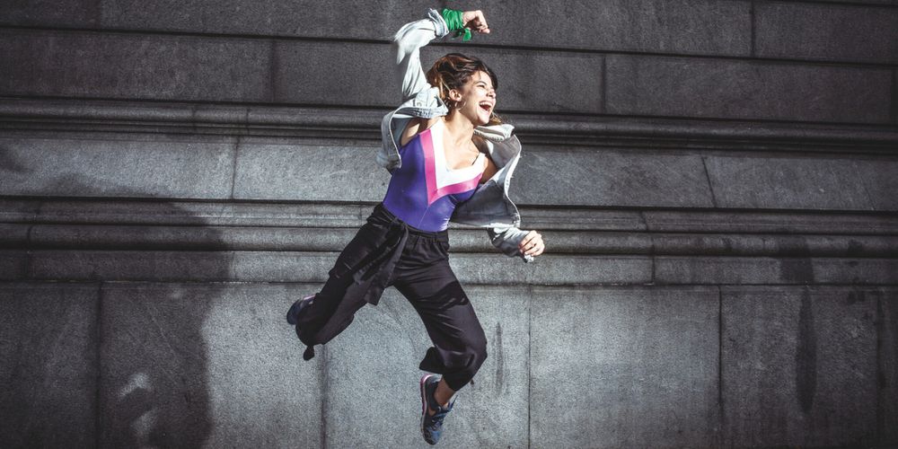 A young woman wearing a green scarf around her wrist jumps into the air, she looks defiant.
