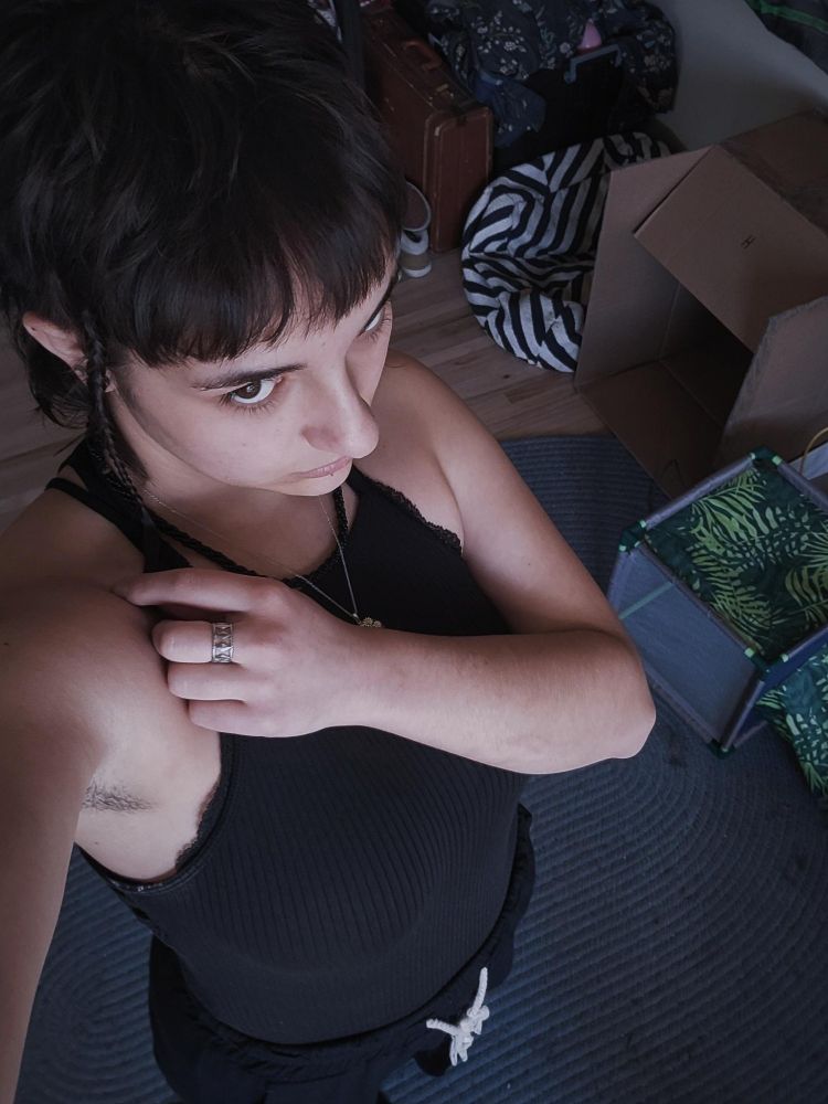 Goth nonbinary babe taking a selfie from above, staring up at the camera and not smiling. Wearing all black and has dark brown hair with bangs.