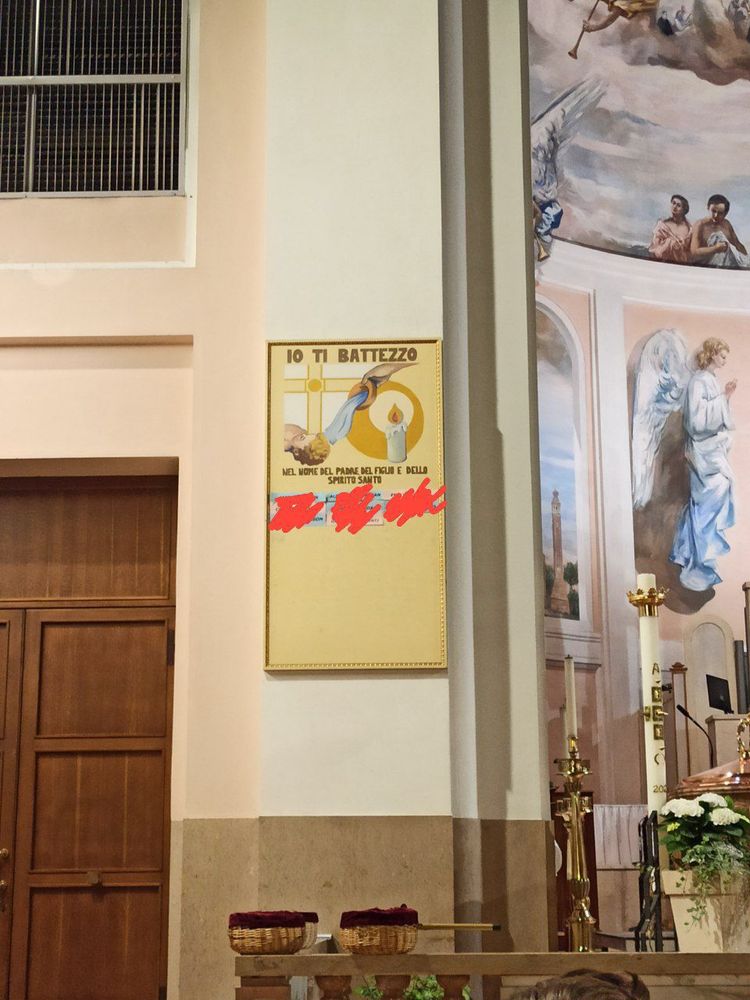 Un pannello con scritto "io ti battezzo nel nome del padre del figlio e dello spirito santo" con dei nomi e cognomi messi su cartoncini azzurro o rosa