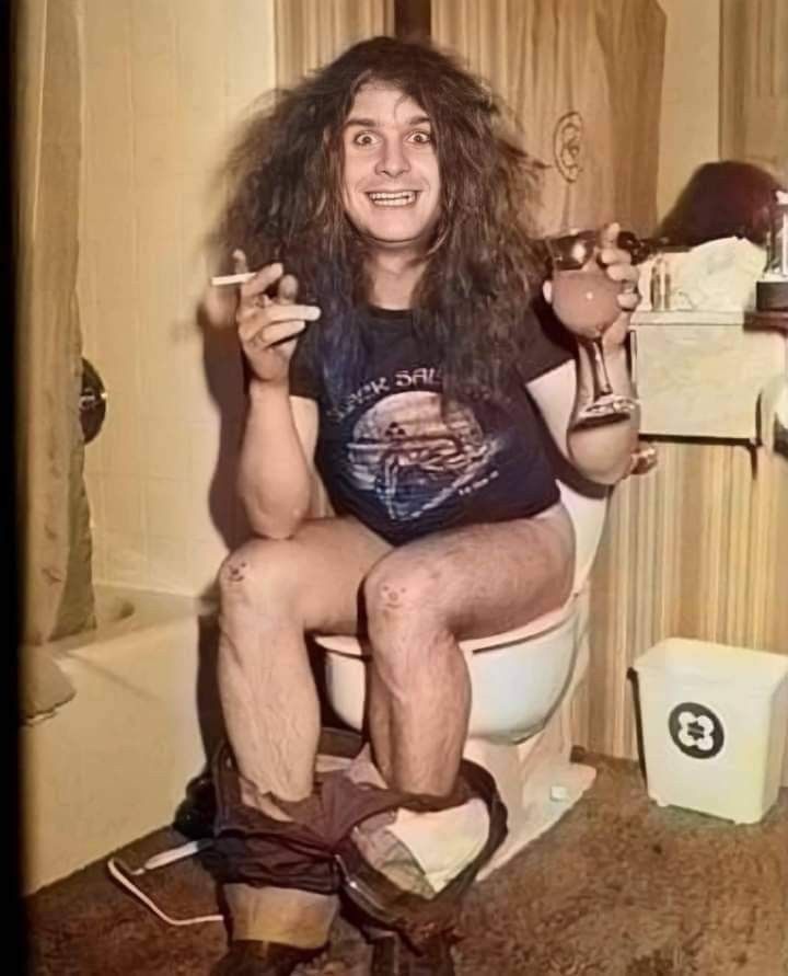 Ozzy Osbourne with a wild mane of hair sits on a toilet with his pants by his ankles. He's got a joint in one hand and a wine glass in another and looks high as hell, eyes bulged. On his knees are drawn two faces. 