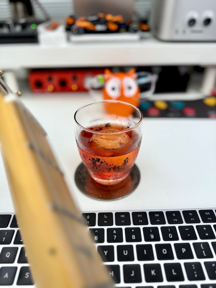 A Mickey Mouse tumbler containing a negroni and an ice sphere. It's on a white desk near a computer keyboard. There's an out-of-focus guitar neck in the foreground and assorted desk nonsense in the background, also out of focus.