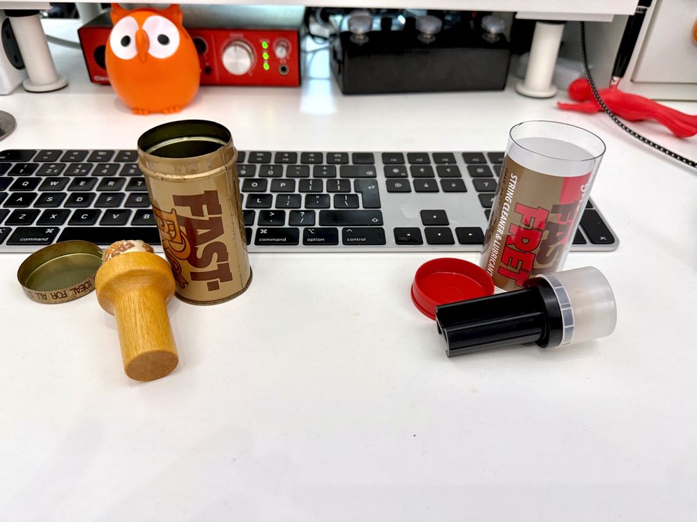 A white desk with desk things. There are also two similarly sized cylindrical containers, both with “Fast Fret String Cleaner and Lubricant” printed on them. The left-hand one is a tin, slightly battered and scratched. Beside it is a small wooden handle with the magic applicator at its end. On the right is a plastic tube. The handle of the applicator is also plastic.