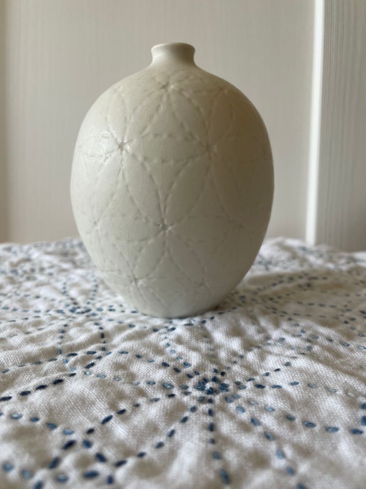 Small white rounded bud vase with all over flower geometric design. Vase is sitting on top of a white cloth sashiko-embroidered in blue with the same design as the vase.