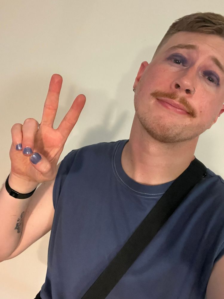Selfie of me giving a peace sign, of course. I’ve got a purple tank top on with purple nail polish and purple eye shadow on. I’ve got a ginger mustache growing in. 