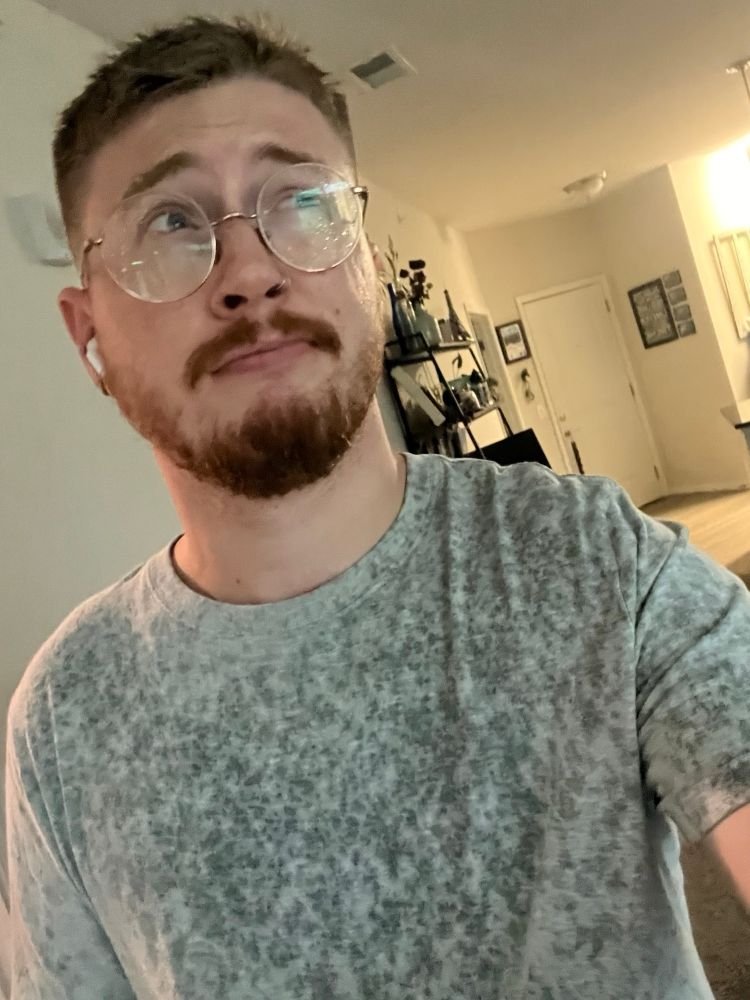 Selfie of me wearing a gray t shirt that is covered in markings from rain drops. I’ve got glasses on and making a facial expression like “ugh, that sucked” 