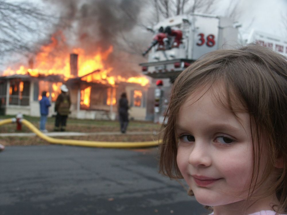 A popular internet meme of a little girl standing in front of a house on fire.  She is looking towards the camera and slightly smiling.
