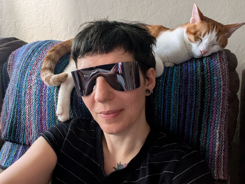 Lia is wearing a pair of shades from the optometrist that wrap around.  Behind her is an orange and white cat draped across the back of a couch.