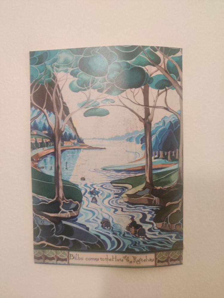 Carte postale patafixée au mur représentant une tapisserie d'Aubusson sur le thème du seigneur des anneaux. La légende brodée dit "Bilbo Comes To the Huts of the Raft Elves" 
