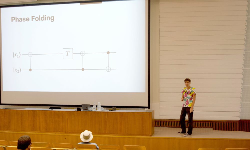 Mark Koch on a stage in an auditorium next to a giant screen showing a very simple quantum circuit.  Mark is wearing a very cool t-shirt.