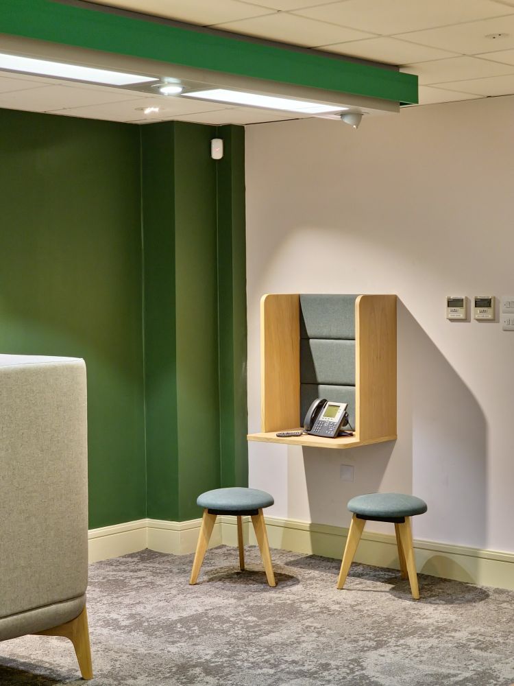 A corner of a bank. One wall is white, the other green while the strip light on the white ceiling is framed in green. Two soft grayish green stools are gathered underneath a tall hanging box shelf that is being used as a booth for a button dial phone with corded handset. Three air conditioning controls are lined up with the top of the booth.