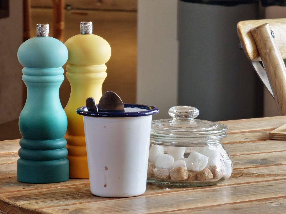 An arrangement of salt and pepper shakers in teal and yellow, next to a white tin cup with blue rim containing teaspoons and a squat glass jar with lid containing sugar cubes. All on a rustic wooden slatted table.
