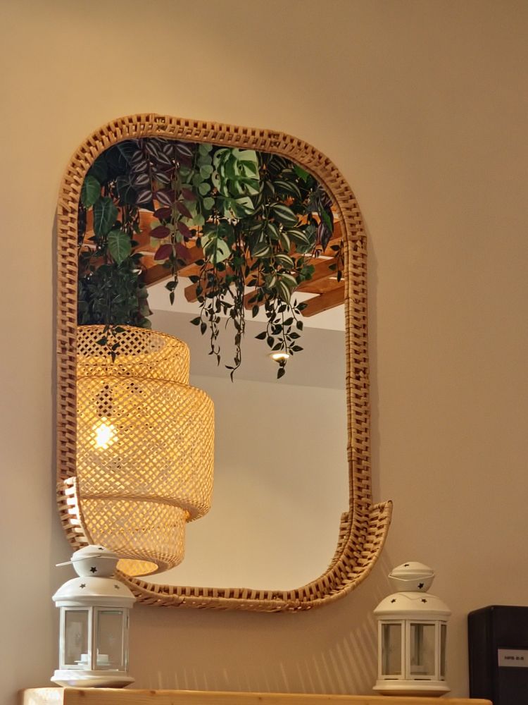 A close-up of a ratten frame mirror on a beige wall. Beneath the mirror is a wooden shelf with white candle lanterns on wither side of the shelf. Reflected in the mirror is the ceiling, from which hangs a wooden frame, dangling plants and a large woven lampshade.