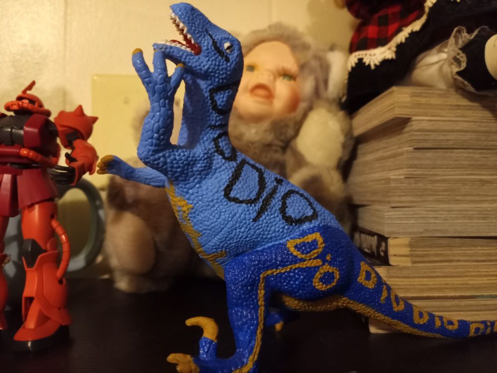 There is a blue Scary Monsters figure from Jojo's Bizarre Adventure in the middle of the picture. Behind the Scary Monster is a cat doll with a human baby face. Who's the real scary monster here?