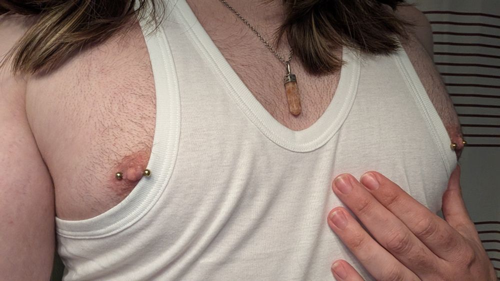 me showing off my chest with a tank top that's pulled too far down, exposing my pierced nipples. my hair is visible draped down my shoulders, as with an orange gem necklace. shot mostly straight.