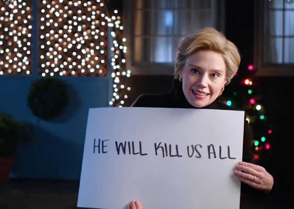 Still image of a SNL sketch with Kate McKinnon as Hillary Clinton holding a placard that says “ HE WILL KILL US ALL” . She is standing outside a persons’ front door mimicking a scene from the movie Love Actually.