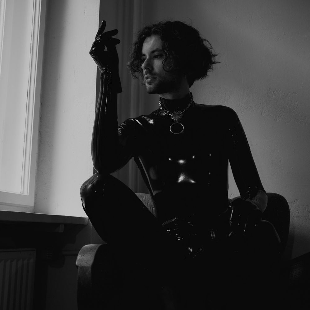 A black and white photo showing Dante squatting on a sofa, wearing full neck to toe black latex as well as latex gloves and an intricate steel necklace with an o ring and a big gem in the centre of it. Dante has medium length wavy hair and looks towards the light coming through the window.