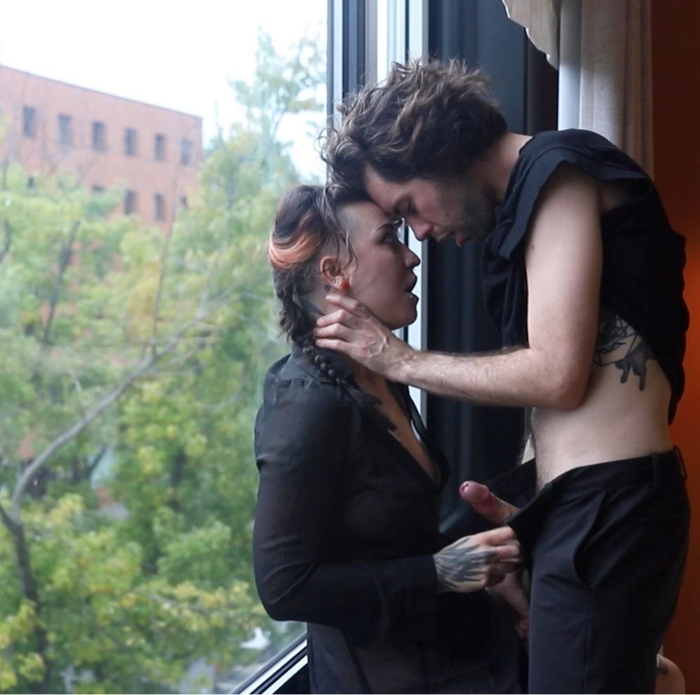 Ursinae leans against a big glass window with Dante standing in front them. Dante chokes Ursinae who opens Dante’s pants, holding on to their hard penis.