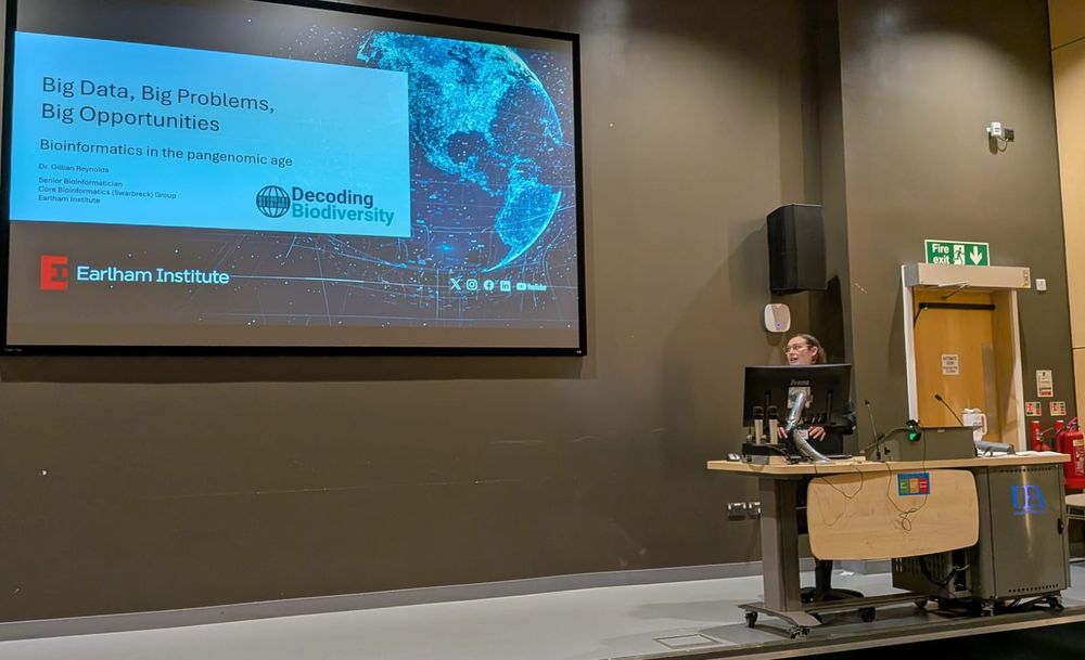 A person presents in a lecture hall beside a large projection screen. The slide shown is titled “Big Data, Big Problems, Big Opportunities: Bioinformatics in the pangenomic age,” delivered by Dr. Gillian Reynolds from the Earlham Institute. The slide features a blue digital globe graphic.