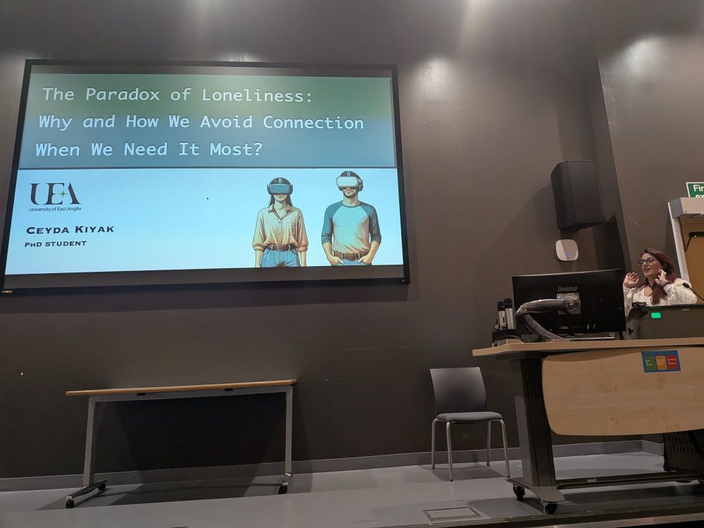 Ceyda Kiyak stands at a lecture podium speaking, with a large presentation slide projected behind her. The slide title reads: “The Paradox of Loneliness: Why and How We Avoid Connection When We Need It Most?” It also shows the University of East Anglia (UEA) logo, the name Ceyda Kiyak, PhD Student, and an illustration of two people wearing virtual reality headsets.